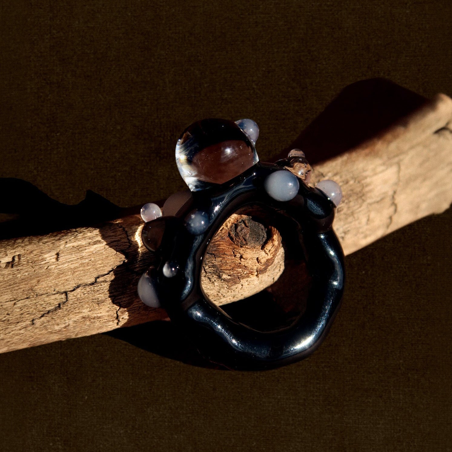 A dark, organic-shaped ring with small white bead accents and a clear dome stone, displayed on a piece of weathered wood against a dark brown background.
